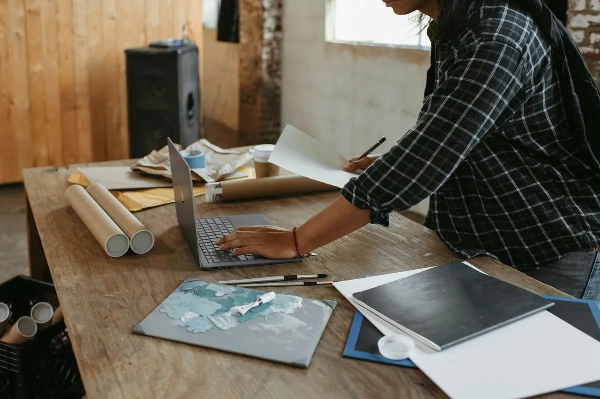 A person in a checked shirt types on a laptop at a large wooden table with drawing tubes and rolled paper.