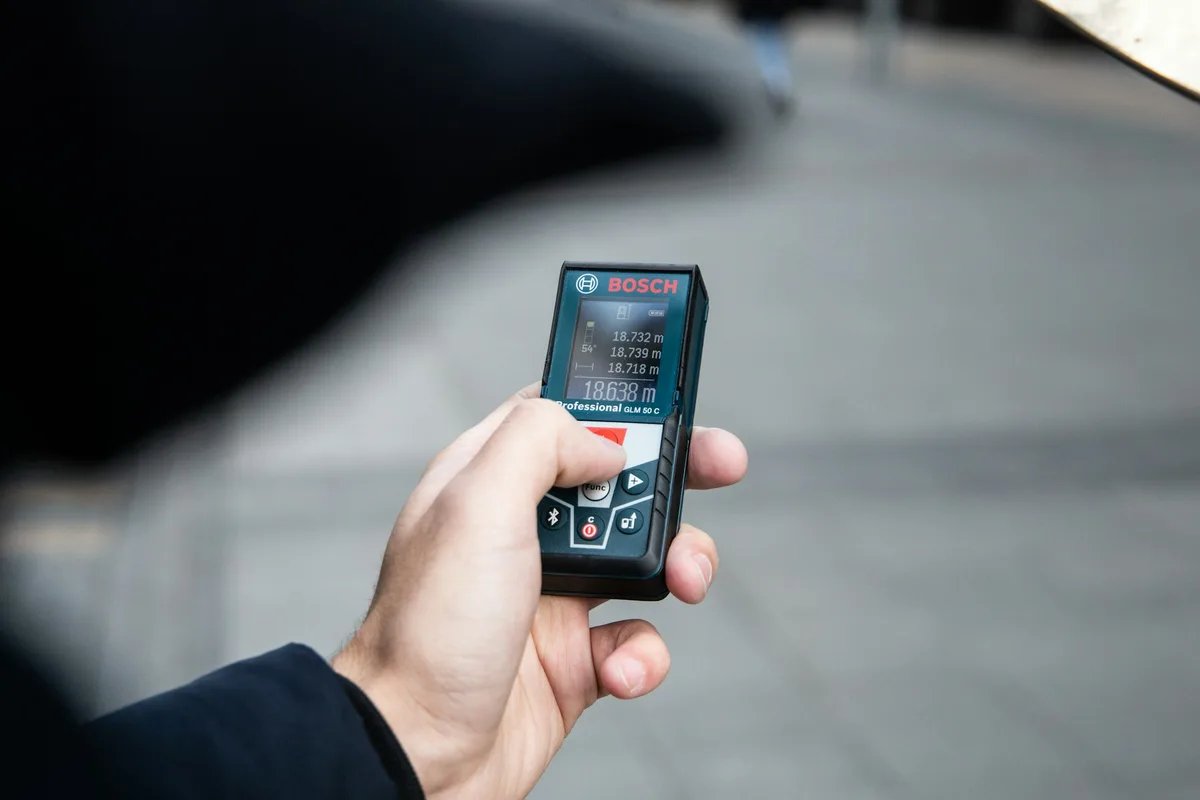 A hand holds a handheld laser distance meter whose screen shows distance readings.