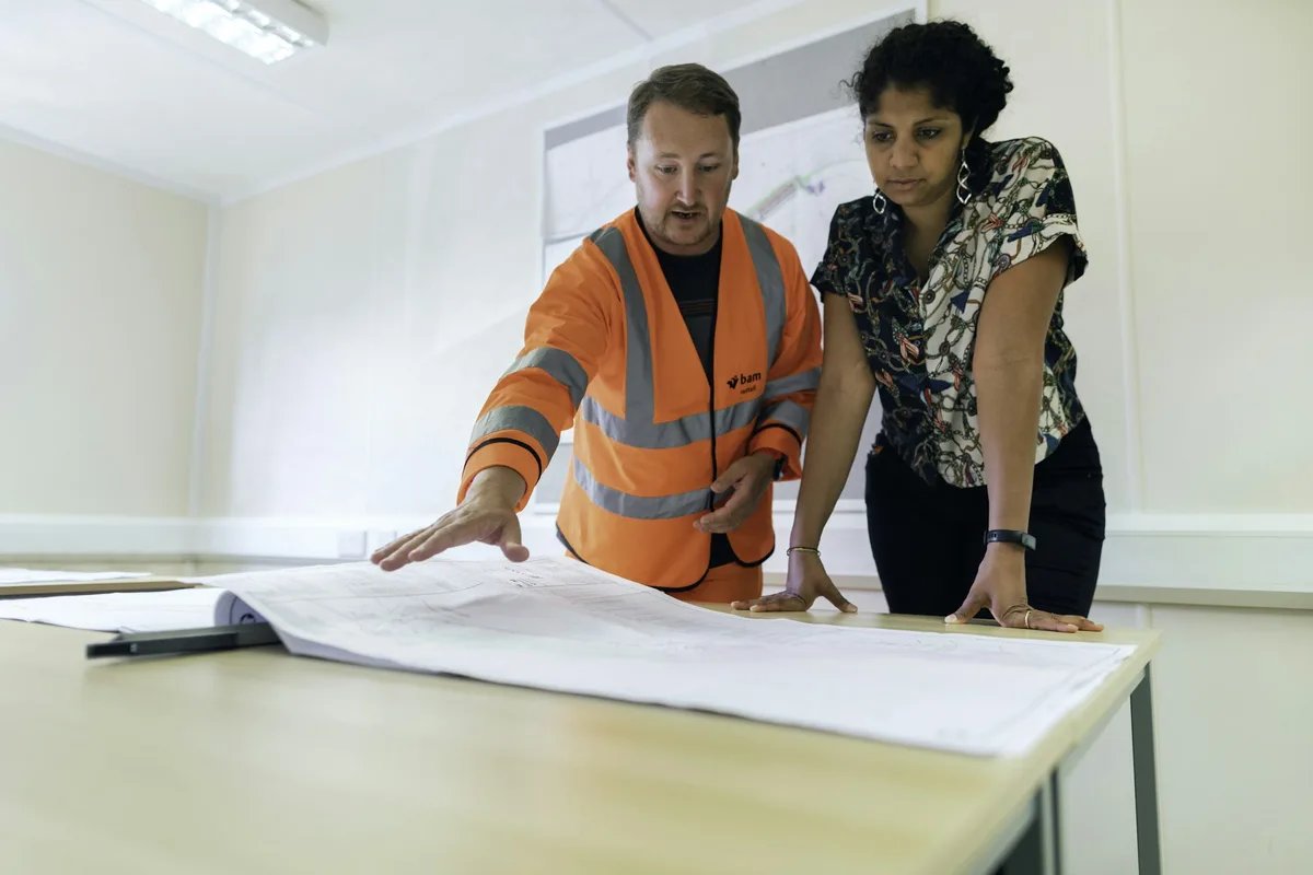 A site worker in an orange hi-vis vest points at a large drawing set on a table while a colleague looks on.