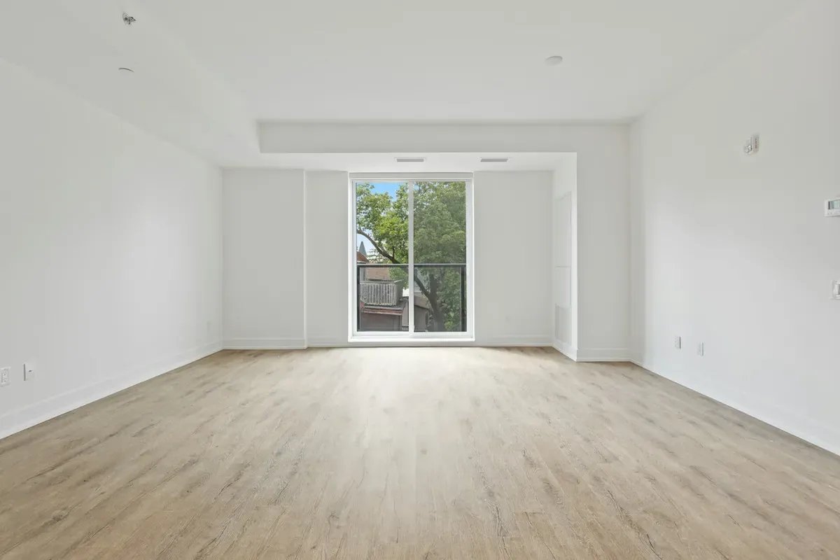An empty white room with a light oak floor and one tall glazed door opening onto a balcony with trees beyond.