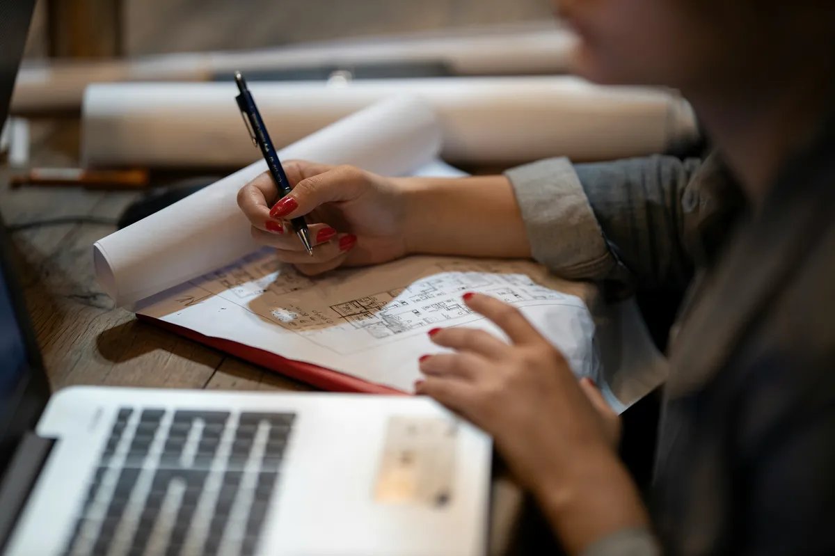 A hand holding a pencil marks up a printed floor plan on a desk beside an open laptop.