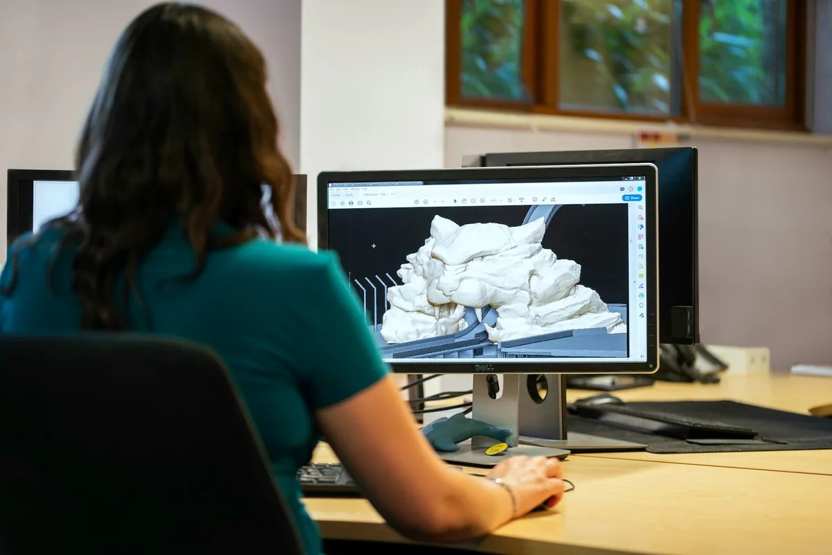 A person sits at a desk and works on a 3D model shown on a computer monitor.