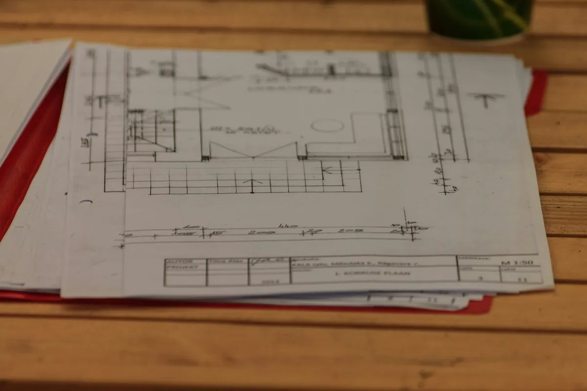 A stack of hand-drawn floor plan sheets on a wooden table, with a title block marked 1:50.