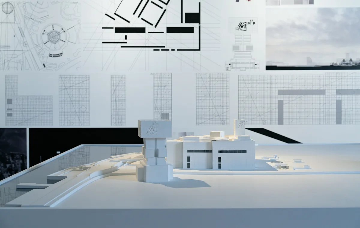 A white massing model of a tower and a low building in front of a wall of plans and site drawings.