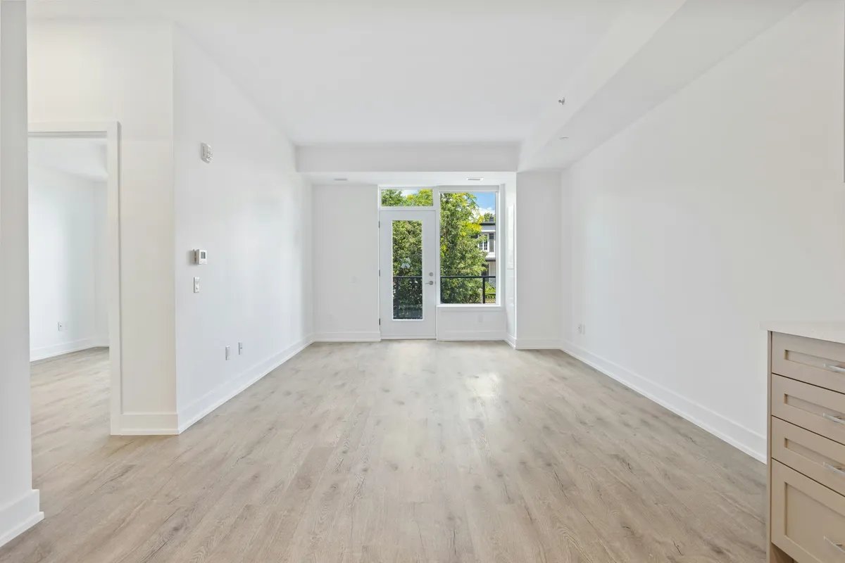 An empty open-plan living room with a bedroom door on the left and a glazed door to a balcony at the far end.