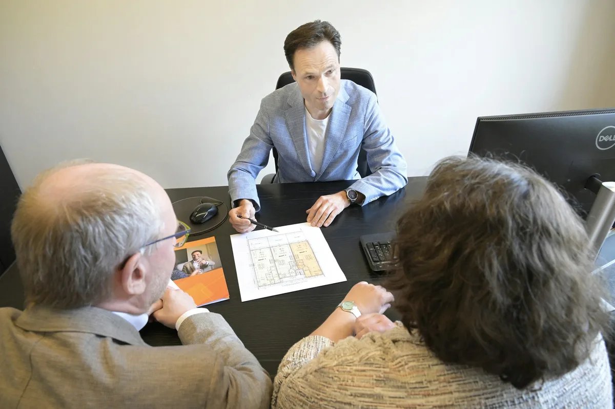 Three people sit at a desk while one of them holds a pen over a printed floor plan during a review.