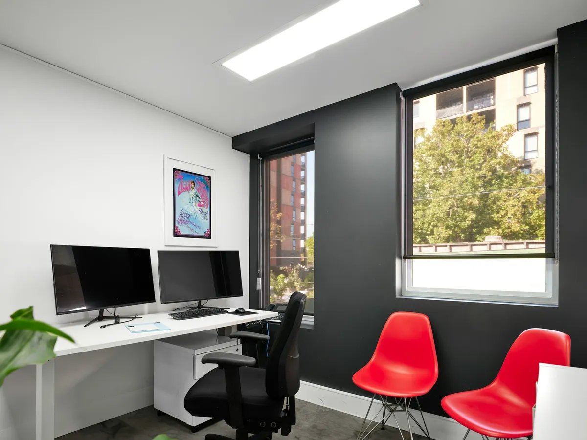 A white office desk with two monitors, a keyboard and a black task chair beside a window onto trees.