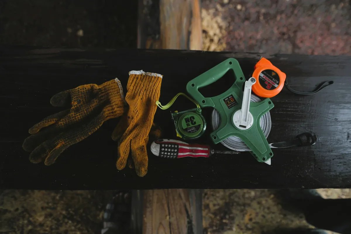 Work gloves, a long open-reel tape and two pocket tape measures lying on a dark timber beam on a building site.