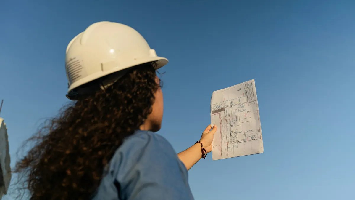 A worker in a white hard hat holds a printed plan up against a clear blue sky, seen from behind.